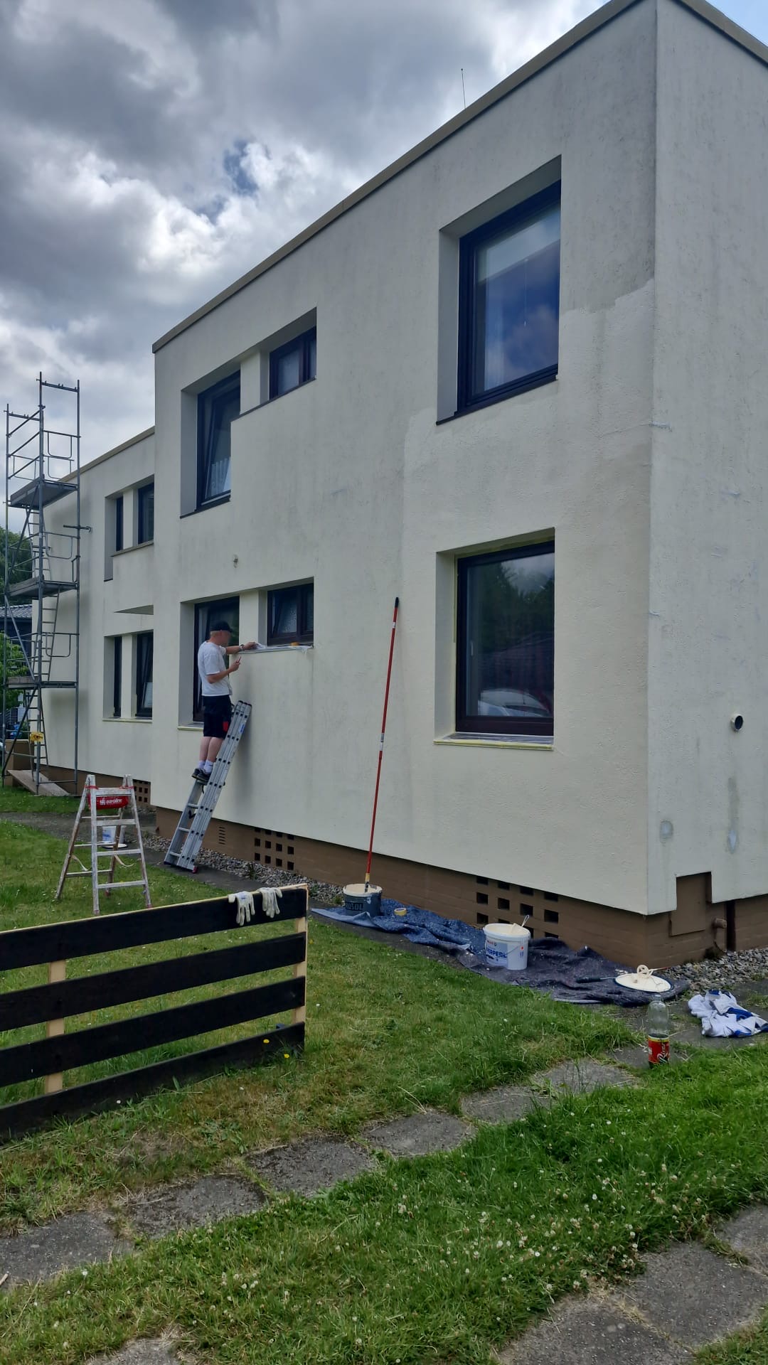 Person auf einer Leiter an einer weißen Hausfassade mit dunklen Fensterrahmen.