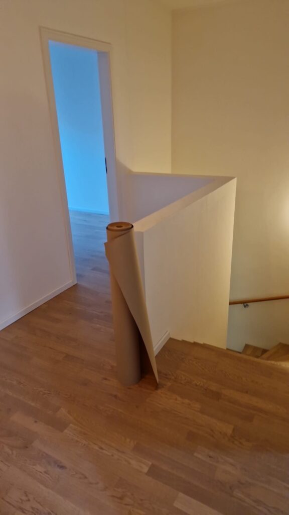 Treppenpodest mit weißen Wänden, Boden in Holzoptik und einer Rolle Abdeckpapier.