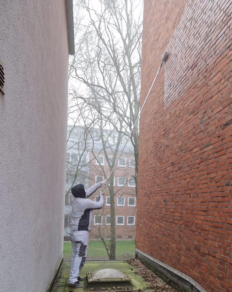 Person mit einer Farbrolle an langer Stange vor einer roten Ziegelfassade.