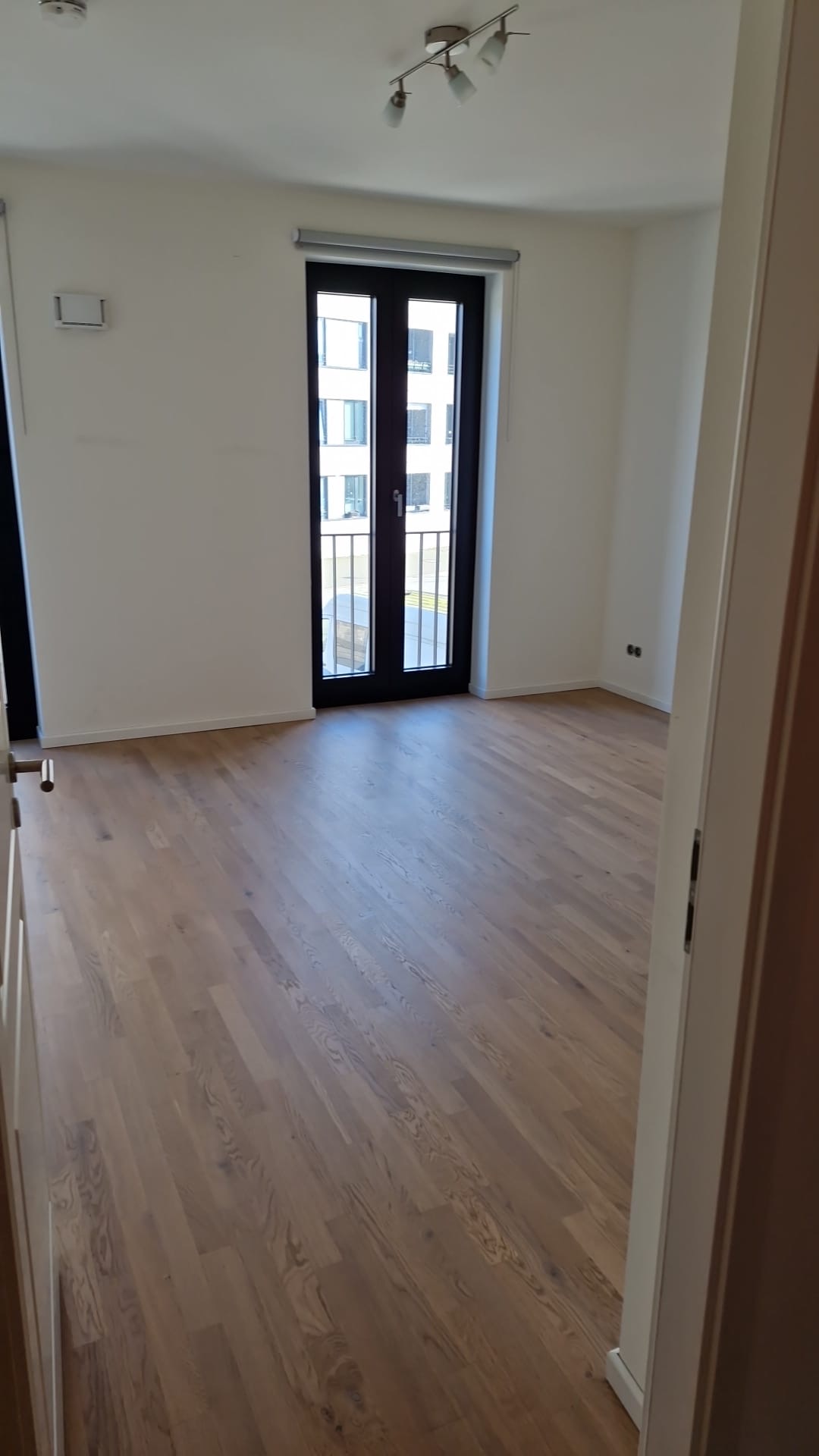 Heller Raum mit weißen Wänden, dunkler Balkontür und Boden in Holzoptik.