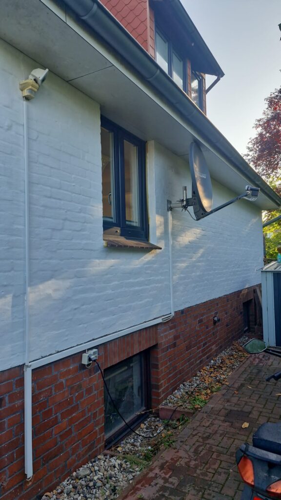 Weiße Ziegelfassade mit dunklem Fenster, Satellitenschüssel und rotem Sockel.
